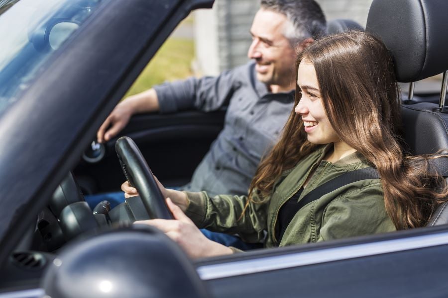 A smiling young woman drives a convertible, with an older smiling man in the passenger seat.